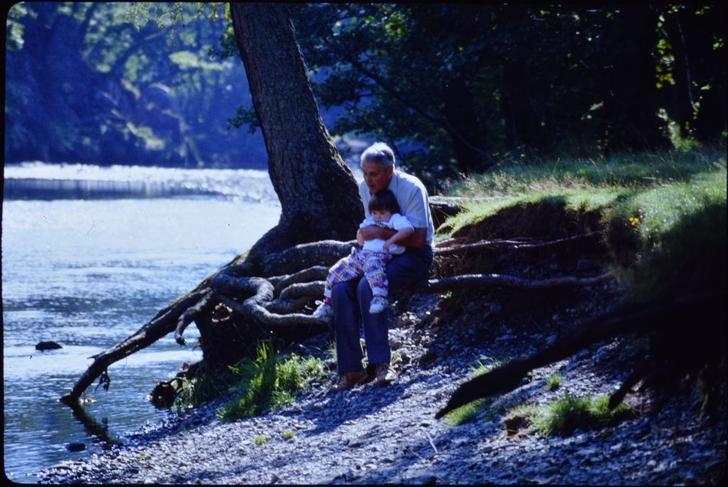 A grandfather holds his grandaughter in his lap at the edge of a river.